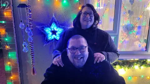 Ollie Conopo/BBC Kerri Frost in a black coat with her hands on Mark's shoulders. He is wearing glasses and a black hoodie. They both stand in front of a front door that is lit up with lots of different coloured lights. 