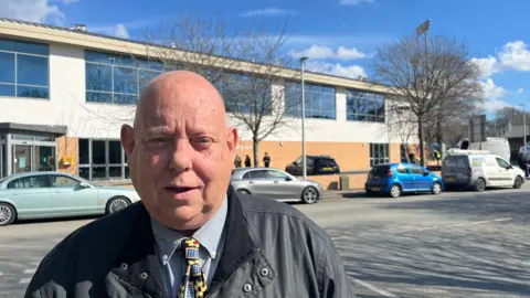 Mark Coker stood to the left looking at the camera. He is wearing a black coat with a blue shirt and yellow, black and blue tie. Behind him is a road with parked cars down one side. There is also a white and brown building on the other side of the road.