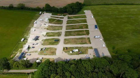 Aerial view of a green field in countryside, which has been concreted over to turn it into a hardstanding grid for caravans. There are four large mobile homes as well as several smaller caravans and other vehicles. This photo was taken in May.