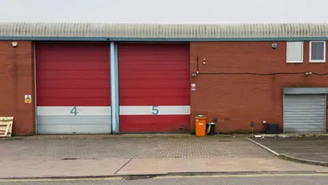 Stephen Menon/BBC A red brick warehouse unit with red shuttered doors with the numbers "4" and "5" and a block paved driveway.