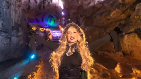 Wookey Hole Caves A woman dressed in black with long flowing blonde hair, smiling to camera and holding her arms aloft in a cave within Wookey Hole cave, which is lit up behind her