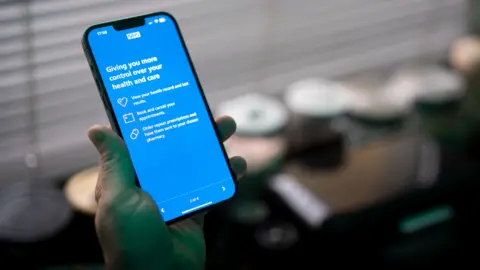A smartphone is held up with an NHS menu visible on the screen.