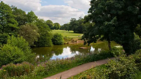 Ian Greig A large green park area with lots of different trees and pathways winding through the open space. There is a large lake in the centre of the shot and a footpath alongside it can be seen in the foreground.
