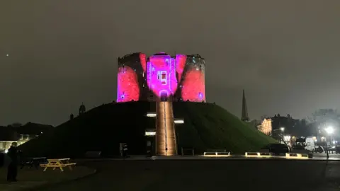 Clifford's Tower in York, a medieval castle sitting on top of a mound of earth with steps leading up the mound to it. The castle is illuminated for a light show.