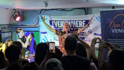PA Media DJ Fatboy Slim raises both arms behind a mixing desk at the front of a small venue as a crowd gathered closely together records the performance on smartphones, with coloured stage lighting and banners promoting the event visible behind.