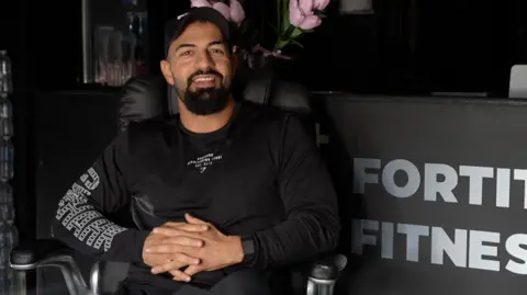 Jamie Niblock/BBC Kamal Mamedov is pictured sitting in a chair in front of a reception desk at his gym. He is wearing a black T-shirt and black trousers, while his hands sit placed in one another in his lap. He is smiling at the camera and is wearing a black cap on his head.