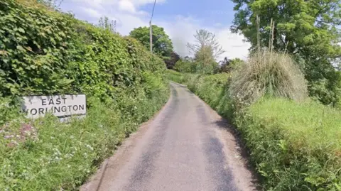 A sign reads EAST WORLINGTON and it is in a hedge next to a single track road. The hedge is lush and has pink and white wild flowers. It is sunny.
