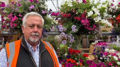 A man with grey hair and a grey beard stands in front of some flowers