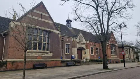 Pete Cooper/BBC Single-storey red brick and stone building - with small windows framed by stone and a stone entrance area. There is a coat of arms over the door and the words "public library" are just visible. There are paving slabs and trees in front of the building.