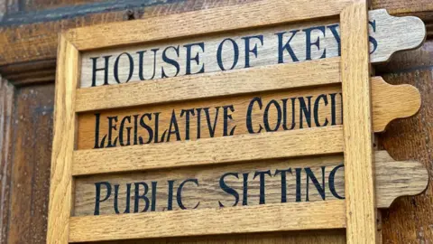 A wooden sign on the door into the parliament building that reads House of Keys, Legislative Council, Public Sitting.