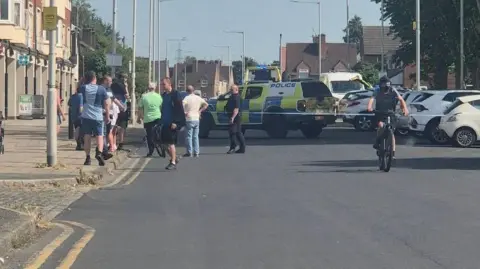 A group of local residents stand in front of a police cordon by a row of terraced townhouse type buildings with shops on the ground floor. Police vehicles and an officer in black uniform are visible behind the cordon. 