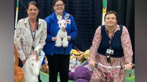 Three women from the school are standing in a sports hall in front of a football goal. One is holding a white and pink stuffed unicorn toy. The next is holding a small white reindeer stuffed animal, and the final woman is straddling a pink and white children's bike