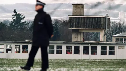 Getty Images A police man in full winter coat and a peaked cap walks in front of brutalist concrete and wood buildings. There is snow on the ground and a lot of snow on hills in the background.