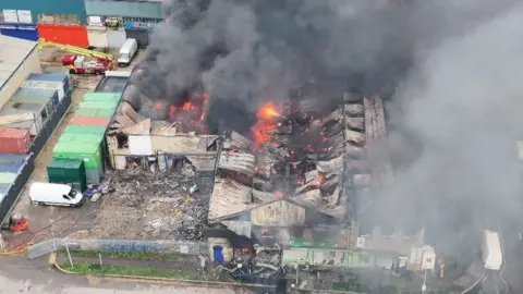 LFB Aerial image shows burnt out building with smoke and flames from it - charred wreckage can be seen beside it
