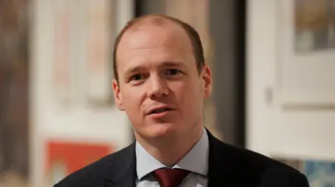 PA Media Gordon Lyons has short dark hair, black suit jacket, white shirt and red tie.