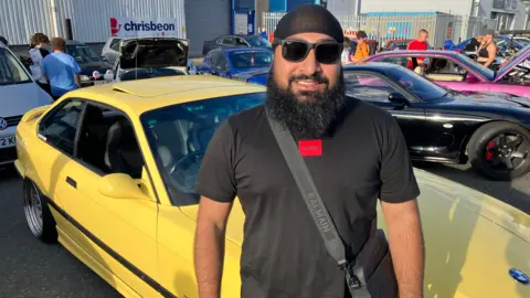 Parmi Singh from Telford standing beside his yellow modified BMW car, He's wearing a black top and sunglasses with a cross the body bag and is smiling into the camera.