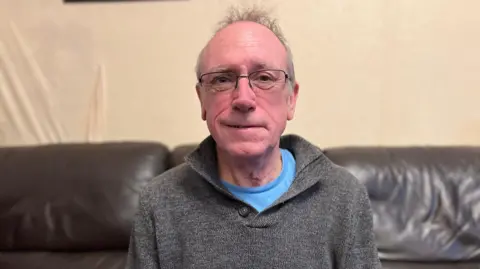 Sandwell and West Birmingham NHS Trust A man sits on a sofa. He has grey hair and glasses. He is wearing a grey jumper with a blue t-shirt underneath. The sofa is made of black leather and has a pink cushion on it.