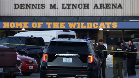 Police officers huddle near a cop car and crime scene tape that is hung up in the parking lot outside an ice skating rink. 
