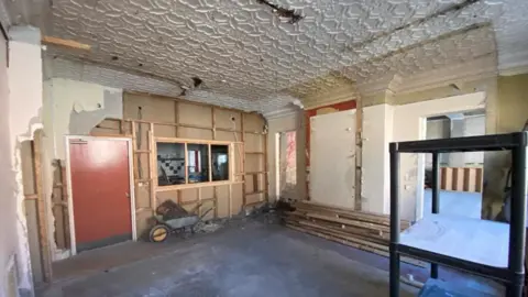 Auction Estates Interior of a room which has been stripped out. One wall appears to be completely plasterboard, a window area overlooks another room, and it has a red door. A wheelbarrow can be seen underneath the window. The two other visible walls have exposed plasterwork. The floor is bare and a metal stand  or shelving can bee seen on the right of the image.