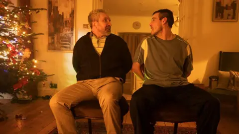 Wildcard Two men are sitting tied to chairs looking at each other. They are in a living room with a Christmas tree in the corner. One man has short grey curly hair and facial hair, and is wearing light coloured trousers, a black zip up and striped top. The other, younger man, has short dark hair and facial hair and is wearing black trousers and a green t-shirt. There are pictures, a clock and curtains behind them in the living room.