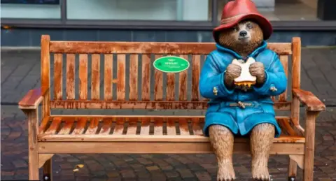 StudioCanal A statue of a Paddington Bear in a red hat and a blue coat holding a sandwich and sitting on a bench