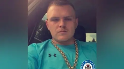 Family A man with short hair and wearing a gold chain and a green T-shirt is sitting in a car.