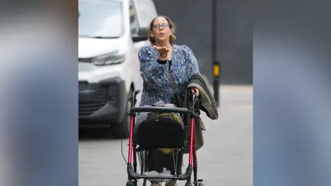 Atidel Boutara Cook arrives outside court with a red Zimmer frame. She is blowing kisses to photographers outside