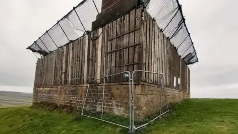A grey fence and wooden scaffolding and black netting in place around a stone structure