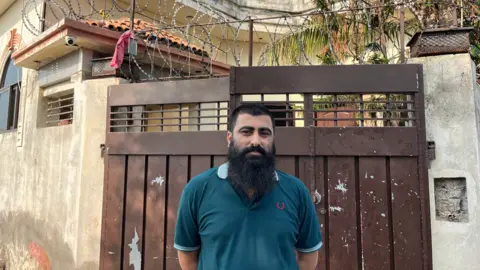 BBC Rasikh Munir is standing in front of the gate to his property where he says he hid Sara Sharif's family. He has short hair, a moustache and a long black beard and is wearing a blue T-shirt.