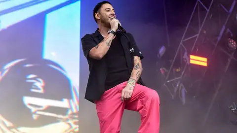 Getty Images A man with dark hair and facial hair on stage singing into a handheld black microphone. He is wearing a black T-shirt, black shirt and pink trousers. He has tattoos on his arms.