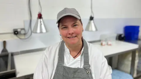 Linda McGibbon, yellowman producer, sitting in her kitchen. Smiling looking at camera. She is wearing a grey baseball cap and grey apron over a white catering jacket. The background is burred and includes heat lamps. 