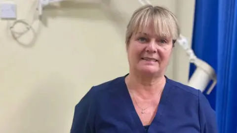 A woman, with blonde hair, wearing dark blue scrubs.