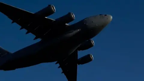 US military plane flies overhead, it is dark grey against a blue sky