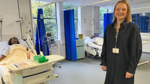 BBC/AMY HOLMES A picture of a woman with blonde hair wearing a dark blue tunic. Behind her are two dummies in hospital beds.