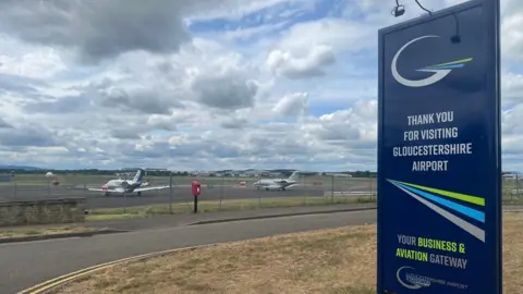 LDRS The exit of Gloucestershire Airport. There is navy sign which reads "Thank you for visiting Gloucestershire Airport your business and aviation gateway" on a scorched grass verge beside a road. On the other side of the road, there is a runway with two small planes sitting on it. The skies above are mostly cloudy.