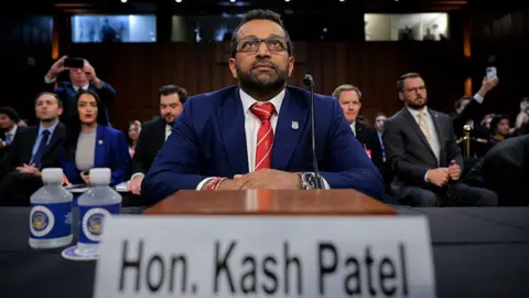 Getty Images FBI Director Kash Patel in a blue suit and red tie standing at a table in the Senate Judiciary Committee, with a small crowd behind him.