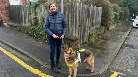 Jessica Bayley/BBC A woman with short grey hair wearing navy jeans with a navy coat and red scarf stood on the edge of the pavement beside her guide dog, a German Shepherd wearing a hi-vis harness, who is looking to the right. There is a fence and bushes behind her.
