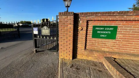 Darren Rozier/BBC The gates of Priory Court Caravan Park. There is a brick wall connecting to an iron fence. 