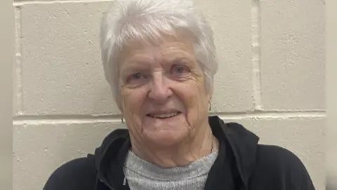 A lady with white hair in a grey jumper wearing a black hoodie. 