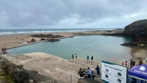 Bude Sea Pool A wide shot of Bude Sea Pool with a sign in the foreground with the title 'Did you know?'. The water is clear and blue-green and there is a beach and the sea behind it.