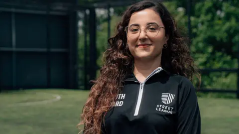 Morgane Bigault / Comic Relief A woman with long curly hair, glasses and a black sports jacket smiles