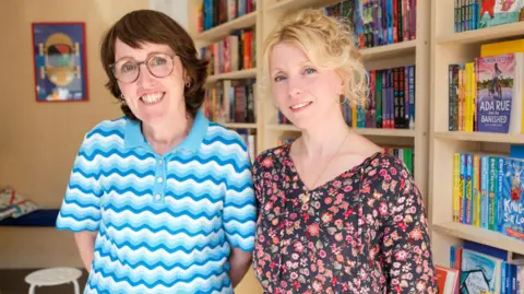 BBC Two women standing in a bookshop. One woman has short brown hair, glasses, and a blue and white t-shirt. The other woman has blonde hair and is wearing a black and red floral dress.