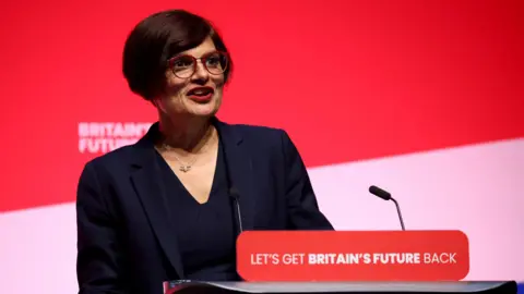 Thangam Debbonaire speaks at a Labour rally, with the podium reading 'Let's Get Britain's Future Back'. She has a red background behind her and wears a black suit.  