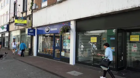 Two empty shop units on King Street in Whitehaven, with a Boots Opticians shop in between them. one on the right has a black frontage with "shop to let" signs in the window. To the left there is another empty unit with a faded wooden front and a "to let" sign.