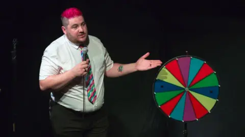 Aaron Weight Aaron Weight, dressed in a white shirt and a colourful tie, stands next to multi-coloured spinning disc, with a microphone in his hand.