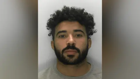 Mugshot of Mohammed Sanana. He has brown eyes and short, dark curly hair and facial hair. He is wearing a grey t-shirt. He is pictured against a plain white background, looking directly at the camera with a blank expression. 