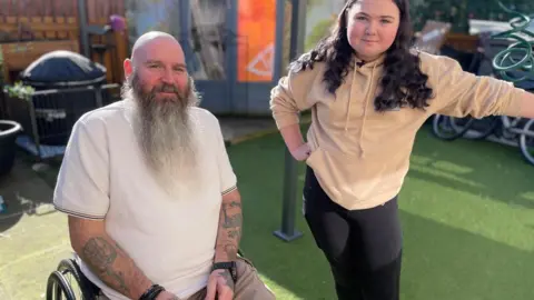 Charly and Mark - Two people are outdoors in a sunny garden area. One person is seated in a wheelchair, wearing a light short-sleeved shirt and has a long beard and visible arm tattoos. The other person is standing beside a metal post, wearing a tan hoodie and dark trousers. Behind them are bicycles, a grill, fencing, and a bright orange panel.