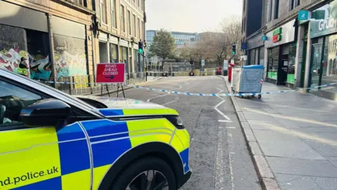 A police car parked across a road, in front of police tape, and a 'road ahead closed' sign with white writing on a red background.