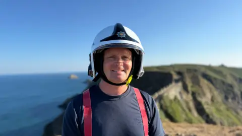 Alex Clarke man in firefighting uniform and a helmet. He is looking at the camera and smiling. Behind him is Sark. The sky is blue. 