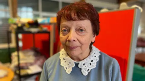 Eileen Wright is the Manager of the Endike Community Care centre. She is stood inside of the centre staring at the camera. She has short auburn hair and is wearing a grey jumper with a white frill collar. Behind her are shelves and piles of paperwork as the staff had to remove it's belongings from the building as it was forced to close.